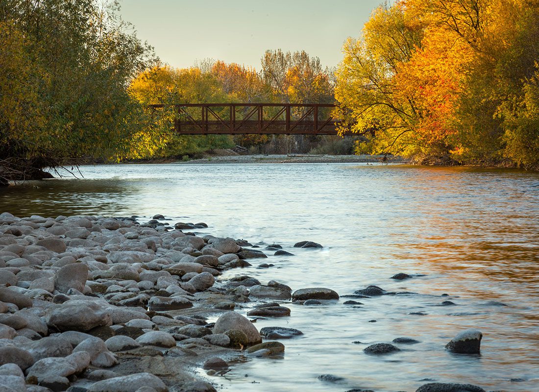 About Our Agency - Scenic View of Rocks on the Edge of a River Surrounded by Colorful Fall Trees with a Steel Walking Bridge Across the Water