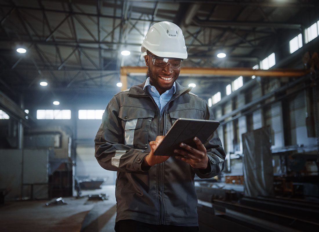 Insurance by Industry - Portrait of a Cheerful Young Male Engineer Standing in a Metal Facility While Using a Tablet