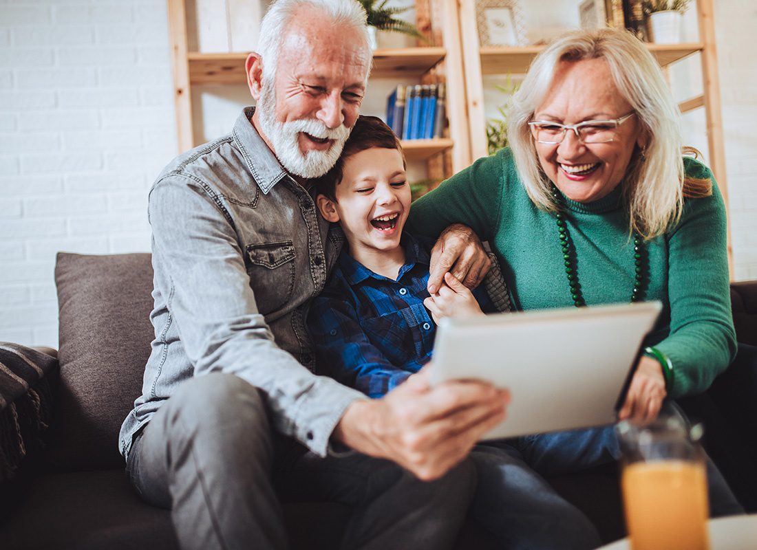 Health Insurance - Portrait of Cheerful Grandparents Sitting on the Sofa in the Living Room with Their Excited Grandson as They Have Fun Watching Videos on a Tablet